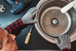 Inside view. Close-up of handsome plumber repairing sink in kitchen. Photo of metalic sink and handyman with screwdriver in gibsonton, fl