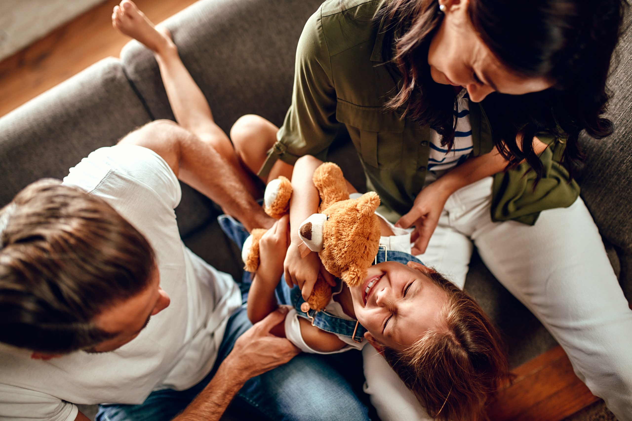 Happy dad and mom with their cute daughter and teddy bear hug, play and have fun sitting on the sofa in the living room at home.