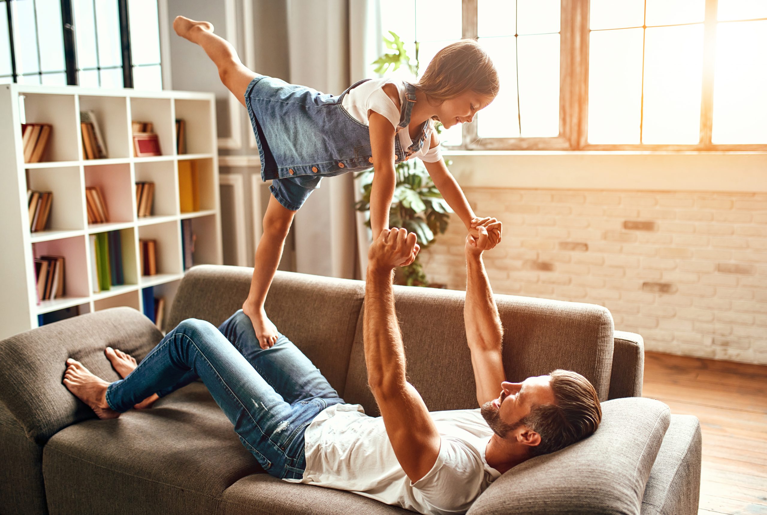 Happy dad fooling around, playing with his daughter on the couch in the living room at home. Happy Father's Day.